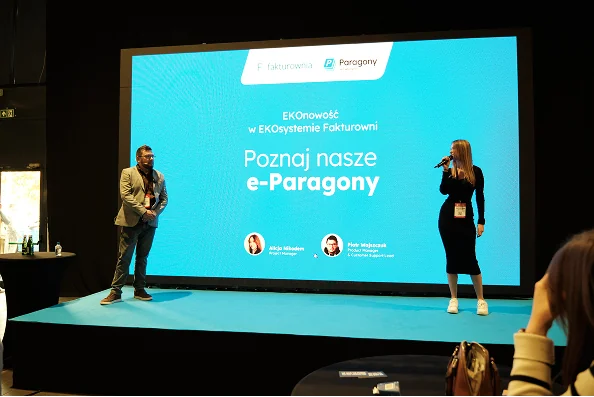 Kobieta z mikrofonem na scenie przed ekranem z napisem Poznaj nasze e-Paragony i logiem Fakturowni