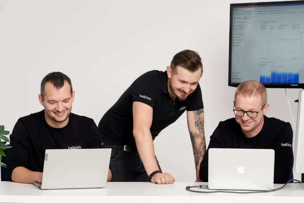 Trzech mężczyzn w koszulkach Tebim przy laptopach, w tle monitor z wykresem